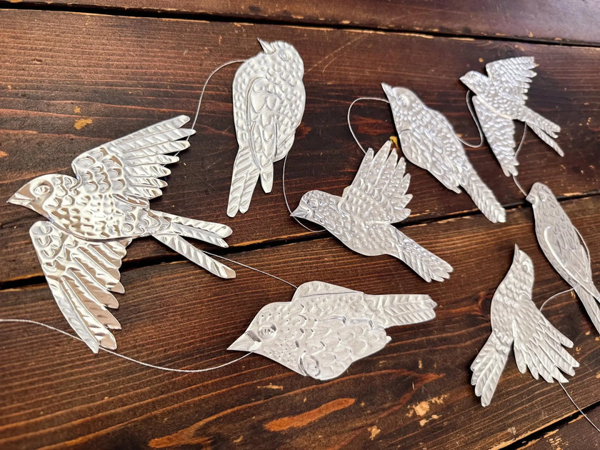 May include: A collection of silver-colored metal bird ornaments with intricate details, some in flight, others at rest. Each bird is suspended by a thin, clear string, creating a delicate, airy feel. The birds are arranged on a dark wooden surface.